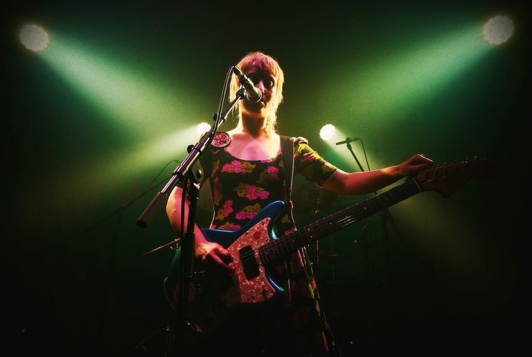 Venus O'Broin from Swapmeet performing on stage in a floral dress, tuning her electric guitar under green spotlights.